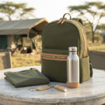 Ein olivgrüner Rucksack mit Lederbesatz steht auf einem rustikalen Holztisch im Freien vor einem Safarizelt. Davor liegen ein zusammengefaltetes olivgrünes T-Shirt und zwei Schlüsselanhänger, einer aus Holz und einer mit einem kleinen Licht. Rechts steht eine Edelstahl-Wasserflasche mit Korkboden.